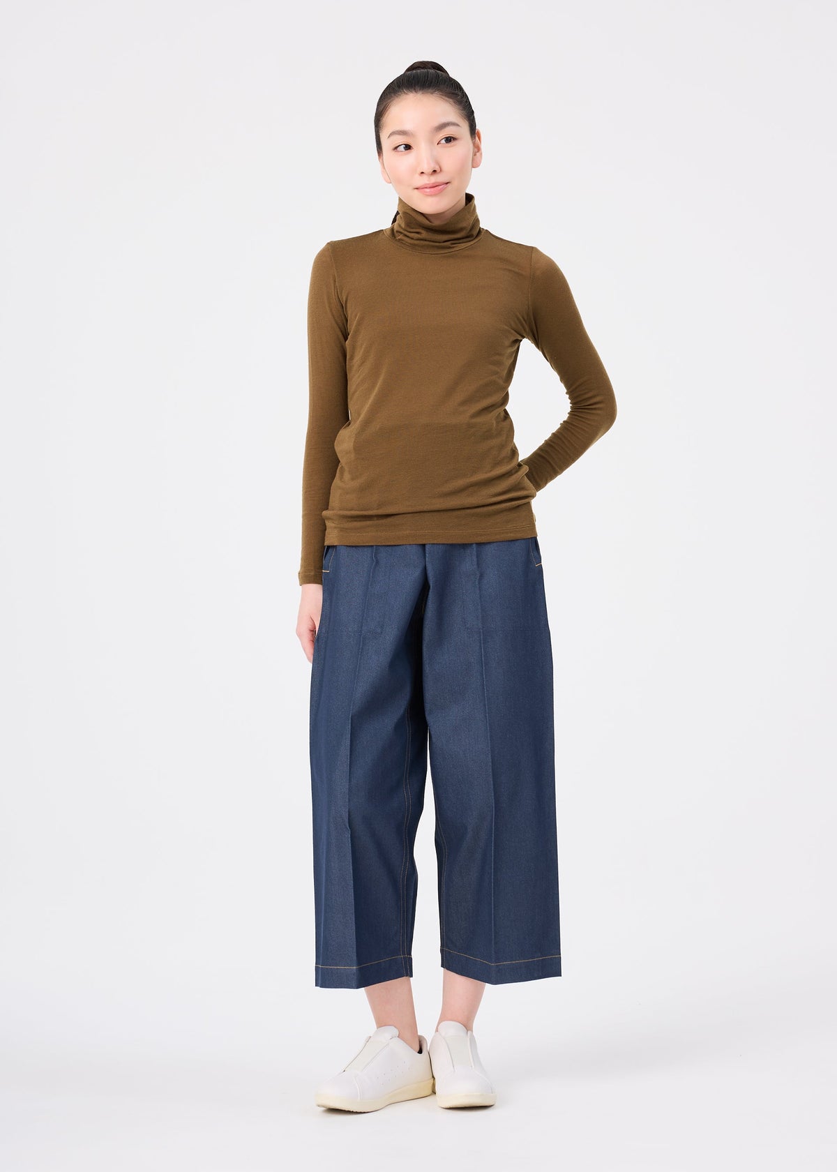 A person stands against a white background, wearing the WOOL MIX JERSEY by me ISSEY MIYAKE, paired with wide-leg blue denim pants and white shoes. Their hands are behind their back, smiling softly to the side. view 8 of 8