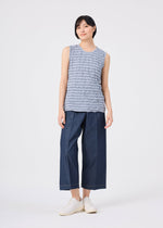 A person with short black hair and a relaxed expression stands against a plain white background, wearing the STRIPE MERINGUE T-SHIRT in light gray by me ISSEY MIYAKE, styled with wide-leg dark blue cropped pants and white slip-on shoes. The sleeveless top features a subtle crushed finish.