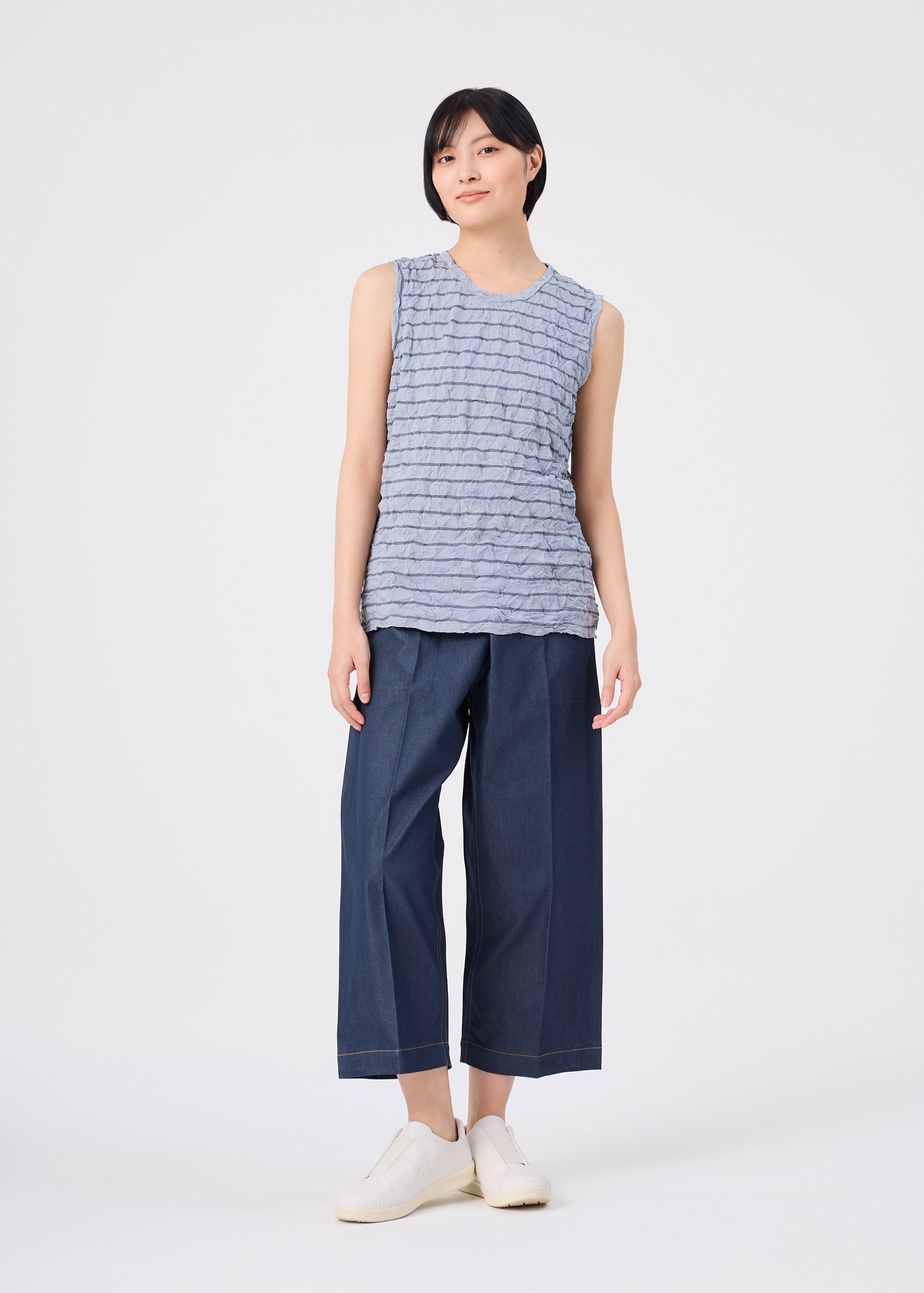 Zoomed view A person with short black hair and a relaxed expression stands against a plain white background, wearing the STRIPE MERINGUE T-SHIRT in light gray by me ISSEY MIYAKE, styled with wide-leg dark blue cropped pants and white slip-on shoes. The sleeveless top features a subtle crushed finish.