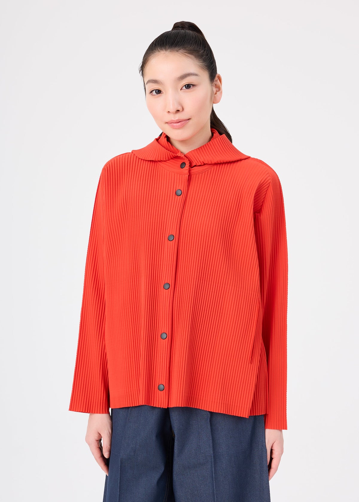 A woman stands against a plain white background, facing the camera with a neutral expression, wearing the FINE KNIT PLEATS COLOR 2 long-sleeve button-up top by me ISSEY MIYAKE in bright orange paired with blue pants. view 7 of 10