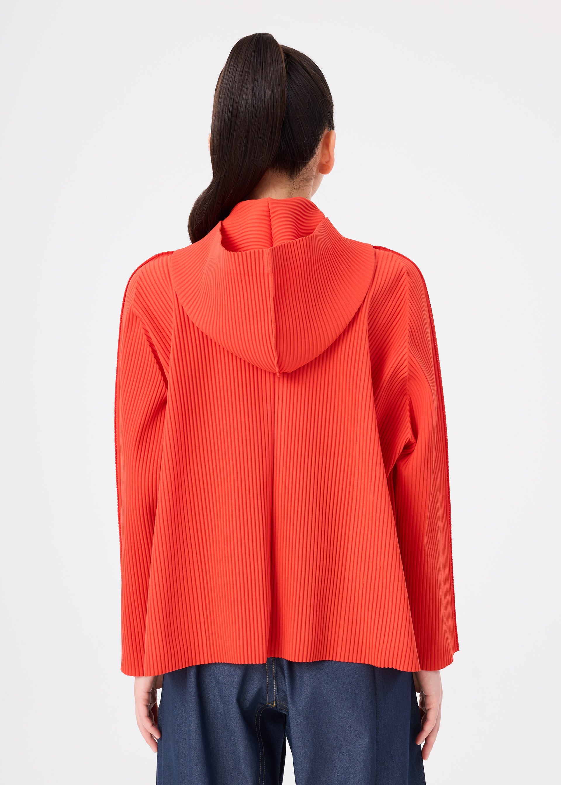 Zoomed view A person with dark hair in a ponytail is shown from the back against a plain white background, wearing a bright red me ISSEY MIYAKE FINE KNIT PLEATS COLOR 2 hoodie and dark blue pants.