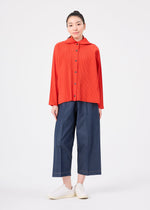 A person stands facing forward against a plain white background, wearing a bright red, long-sleeve button-up top layered over the me ISSEY MIYAKE FINE KNIT PLEATS COLOR 2 hoodie, wide-leg blue cropped pants, and white sneakers.