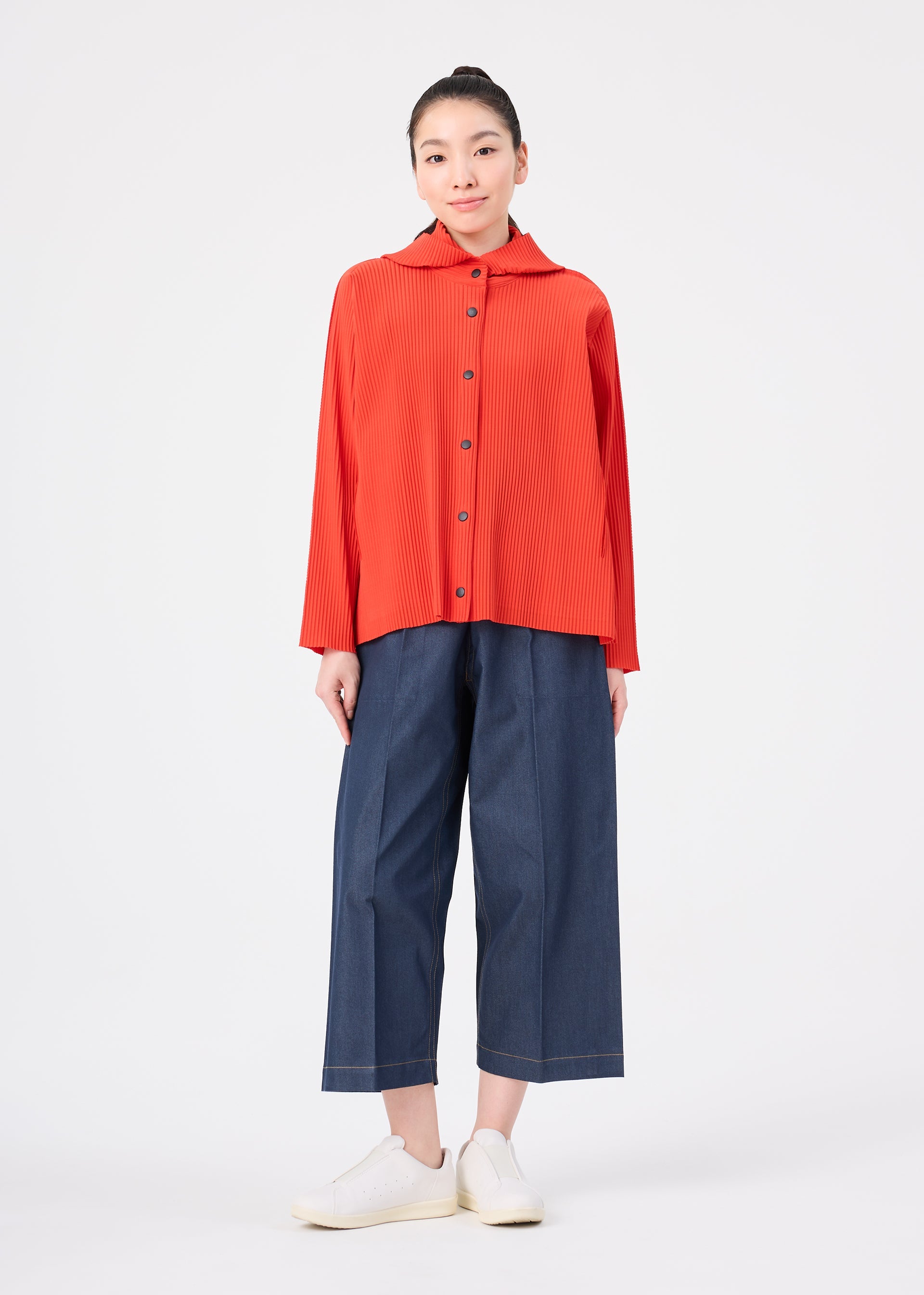 Zoomed view A person stands facing forward against a plain white background, wearing a bright red, long-sleeve button-up top layered over the me ISSEY MIYAKE FINE KNIT PLEATS COLOR 2 hoodie, wide-leg blue cropped pants, and white sneakers.