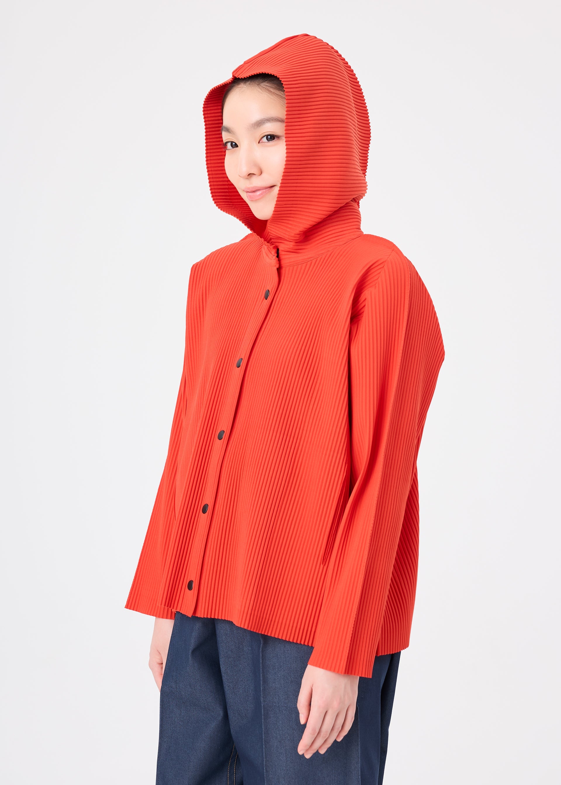 Zoomed view A person wearing the me ISSEY MIYAKE FINE KNIT PLEATS COLOR 2, a vivid orange pleated knit button-up hooded top, paired with dark pants, stands against a plain white background, gazing slightly to the side with a subtle smile.