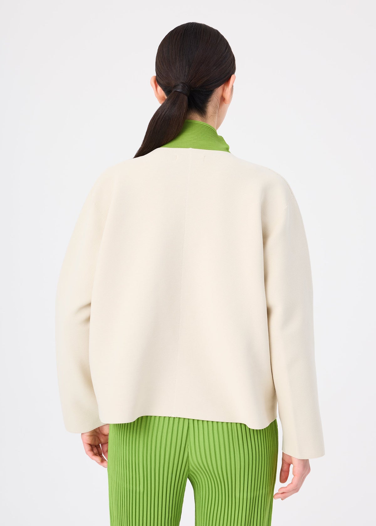 Zoomed view A woman with long dark hair in a low ponytail wears the SOLID KNIT by me ISSEY MIYAKE, layered over a green shirt and green pleated pants, photographed from behind against a plain white background.