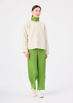 A person stands wearing the SOLID KNIT jacket by me ISSEY MIYAKE over a green turtleneck and green pleated pants with white shoes, against a plain white background.