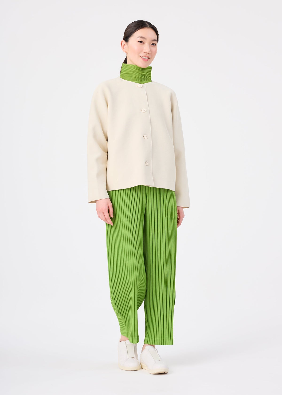 Zoomed view A person stands wearing the SOLID KNIT jacket by me ISSEY MIYAKE over a green turtleneck and green pleated pants with white shoes, against a plain white background.