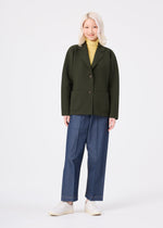 A person with long, light blonde hair smiles in the me ISSEY MIYAKE SOLID KNIT, styled with a dark green blazer, yellow turtleneck, wide-leg blue jeans, and white shoes against a plain white background.