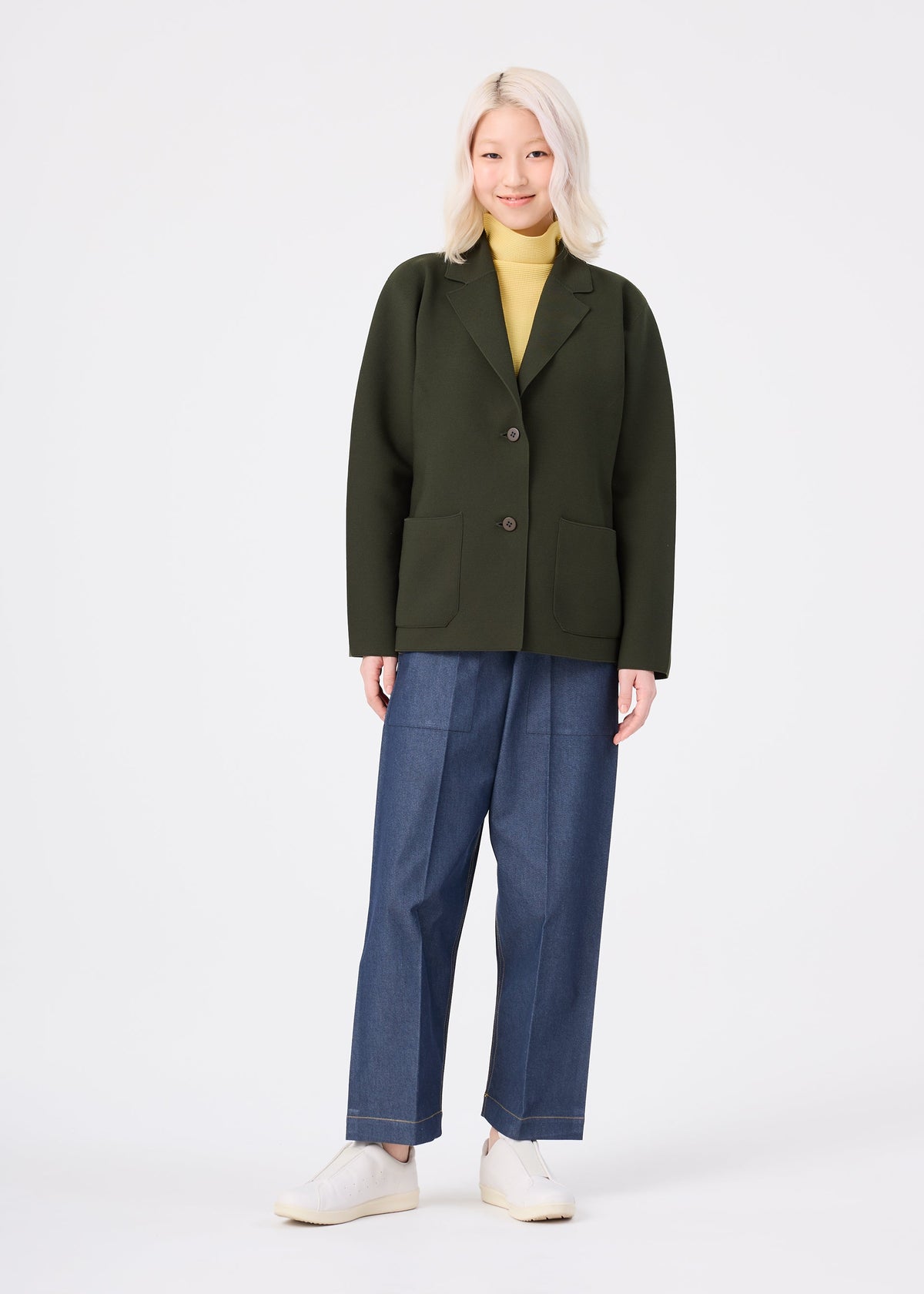 Zoomed view A person with long, light blonde hair smiles in the me ISSEY MIYAKE SOLID KNIT, styled with a dark green blazer, yellow turtleneck, wide-leg blue jeans, and white shoes against a plain white background.
