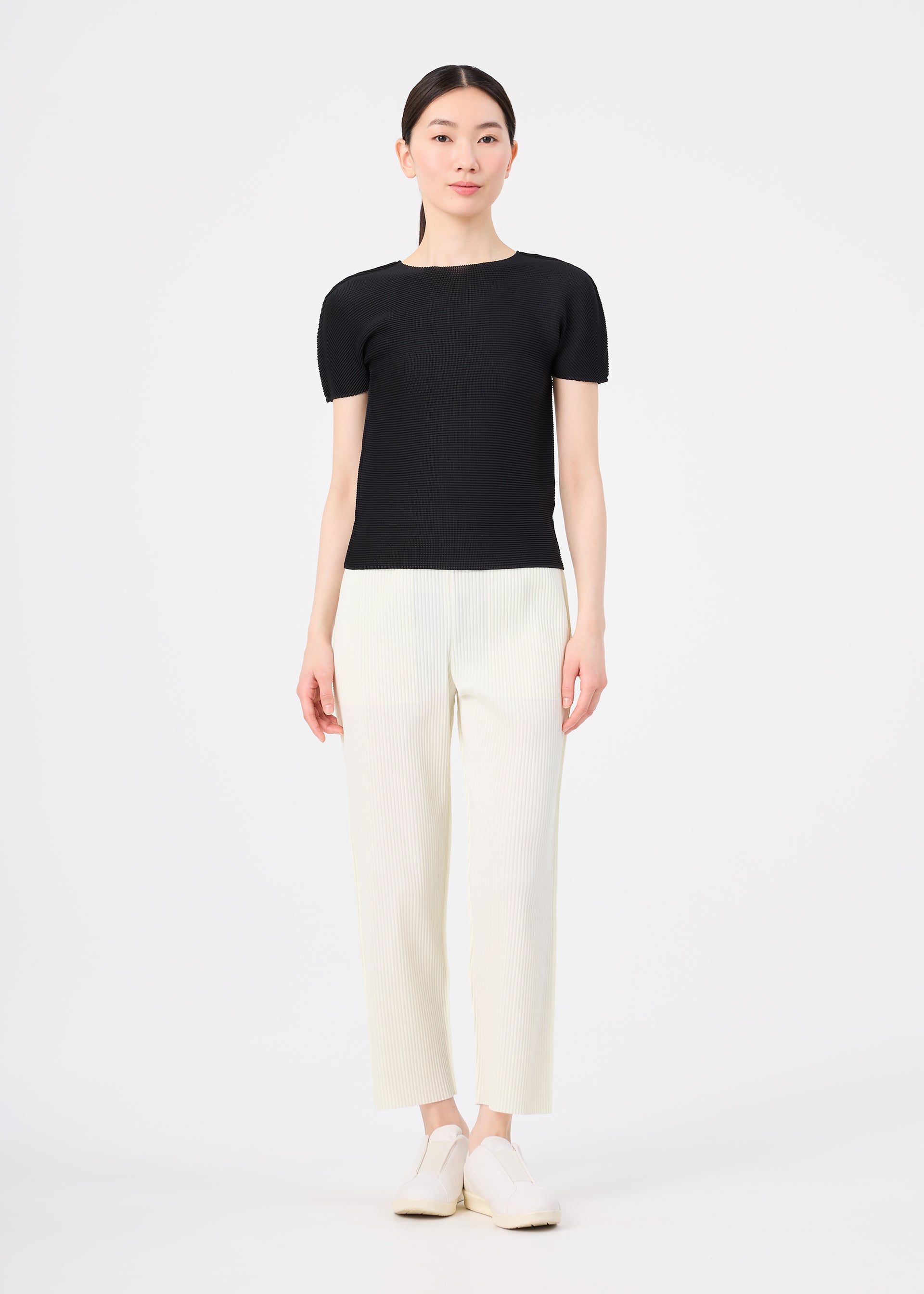 Zoomed view A woman with straight dark hair pulled back and a neutral expression wears the FINE KNIT PLEATS top by me ISSEY MIYAKE, paired with lightweight white straight pants and white shoes, standing against a white background.