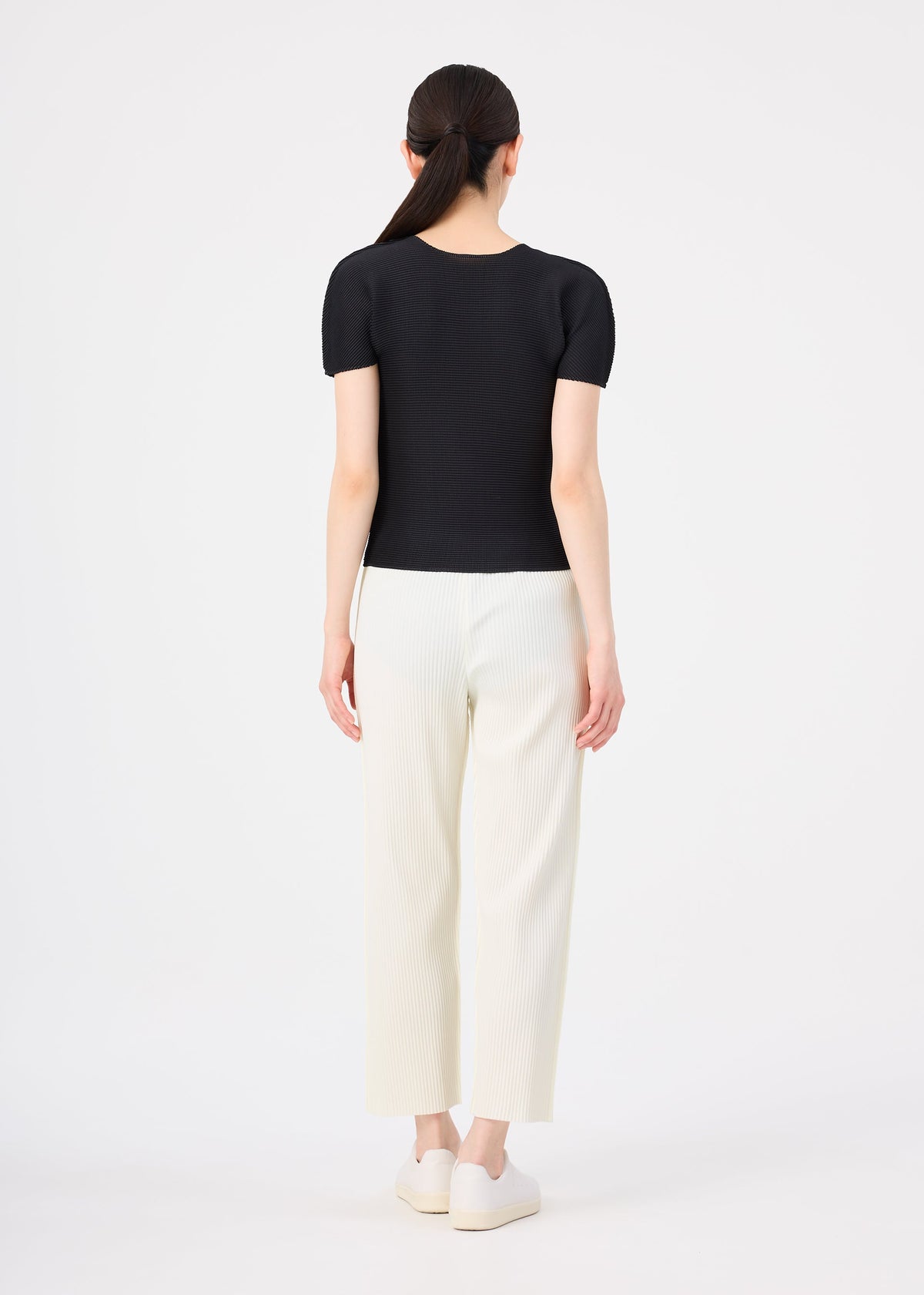 A woman faces away in a fitted black FINE KNIT PLEATS top by me ISSEY MIYAKE, lightweight white pants, and platform shoes. Her straight dark hair is in a low ponytail against a plain white background. view 5 of 5