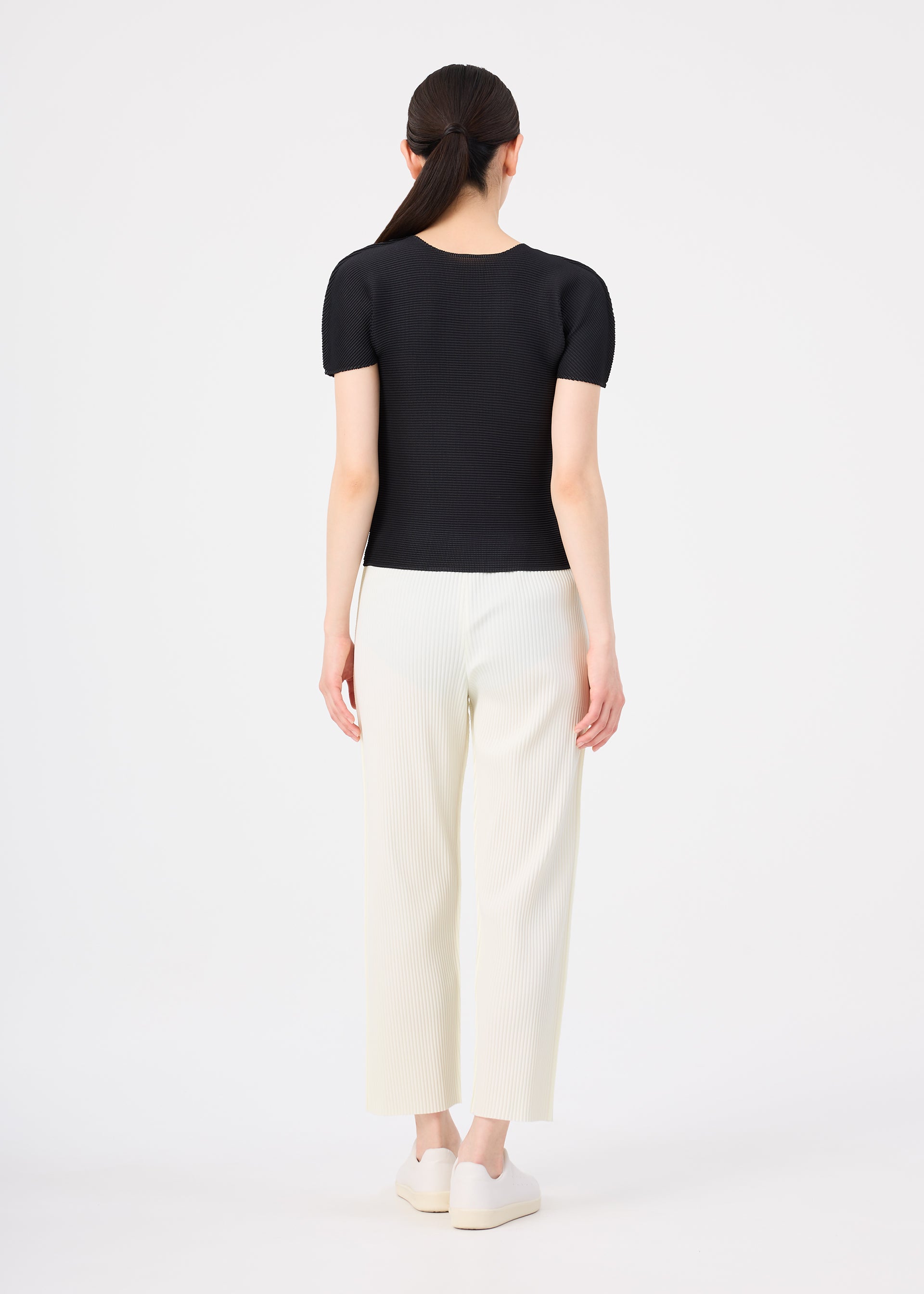 Zoomed view A woman faces away in a fitted black FINE KNIT PLEATS top by me ISSEY MIYAKE, lightweight white pants, and platform shoes. Her straight dark hair is in a low ponytail against a plain white background.