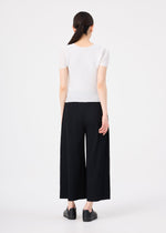 A woman with long dark hair in a low ponytail stands facing away, wearing me ISSEY MIYAKE FINE KNIT PLEATS wide pants, a short-sleeved white top, and black shoes against a plain white background.
