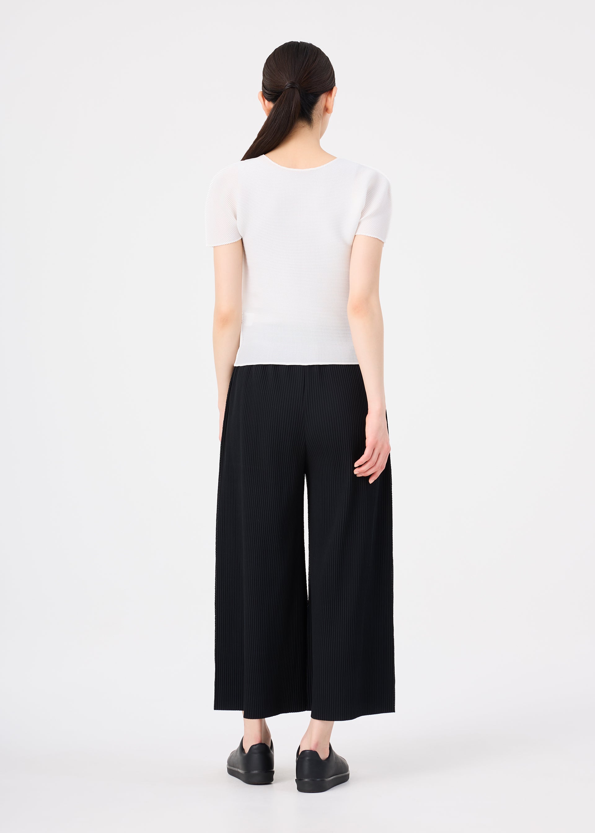 Zoomed view A woman with long dark hair in a low ponytail stands facing away, wearing me ISSEY MIYAKE FINE KNIT PLEATS wide pants, a short-sleeved white top, and black shoes against a plain white background.