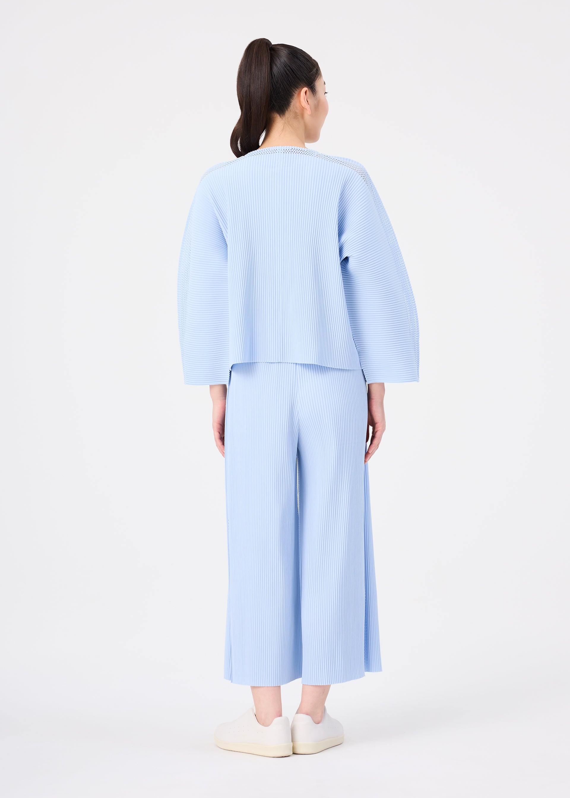 Zoomed view A woman with a high ponytail faces away, wearing the MESH PLEATS lightweight knit pleated top and wide pants by me ISSEY MIYAKE, paired with white slip-on shoes against a plain white background.