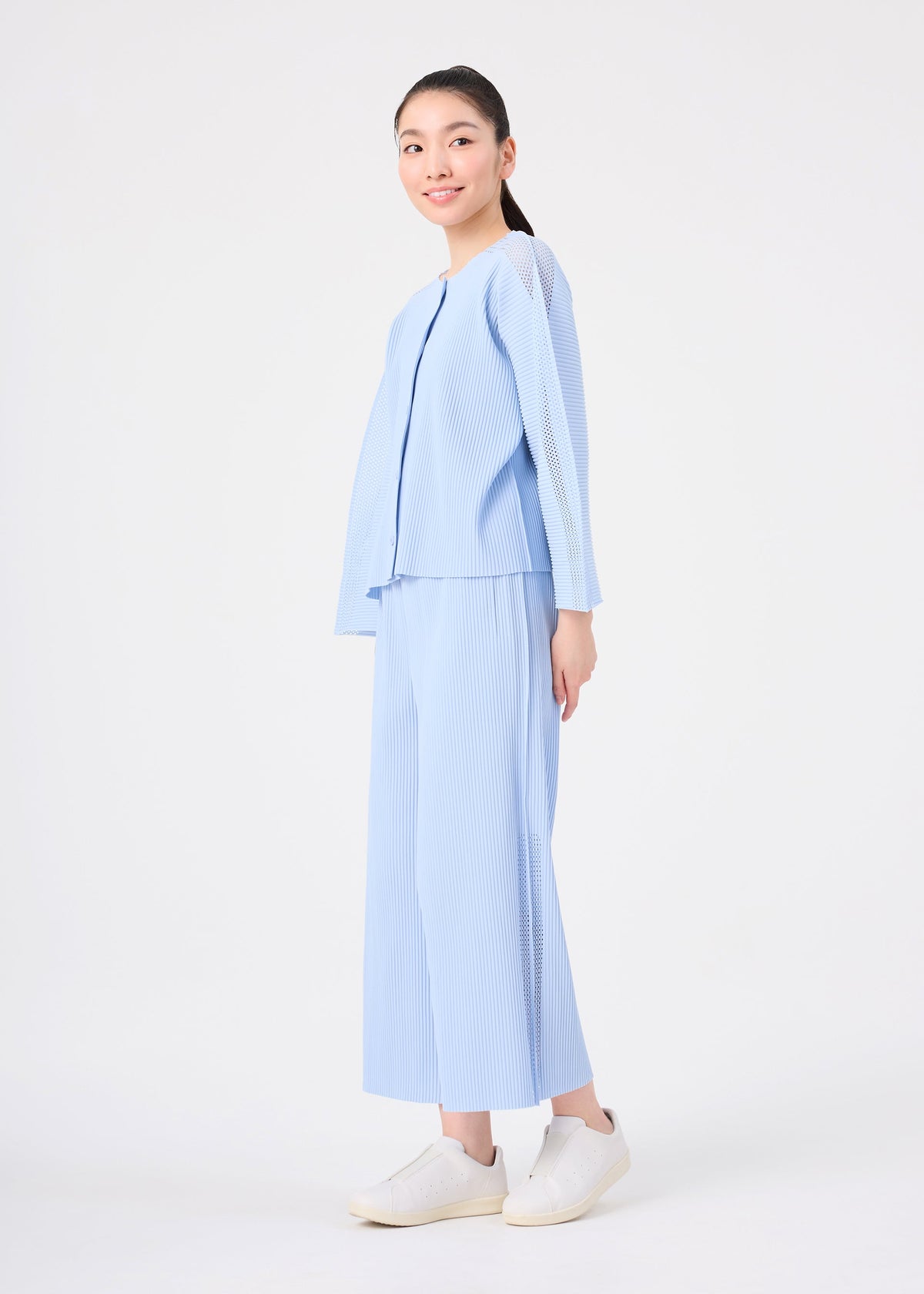 A young woman smiles at the camera against a white background, dressed in the MESH PLEATS set from me ISSEY MIYAKE—light blue, pinstriped top and matching wide pants—paired with white sneakers. view 7 of 8