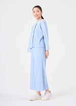 A young woman smiles at the camera against a white background, dressed in the MESH PLEATS set from me ISSEY MIYAKE—light blue, pinstriped top and matching wide pants—paired with white sneakers.