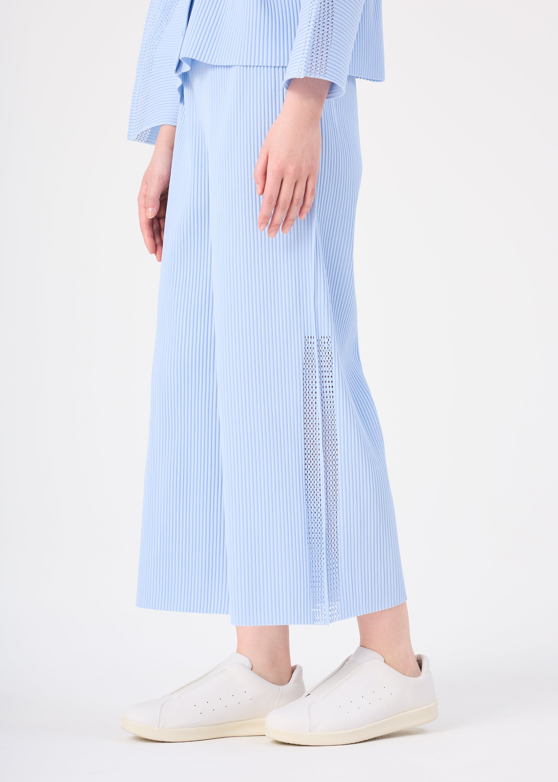 Zoomed view A person wears me ISSEY MIYAKE's MESH PLEATS light blue wide-legged pants featuring fine knit pleats and mesh panels near the hem, paired with a matching top and white slip-on sneakers. The background is plain white.