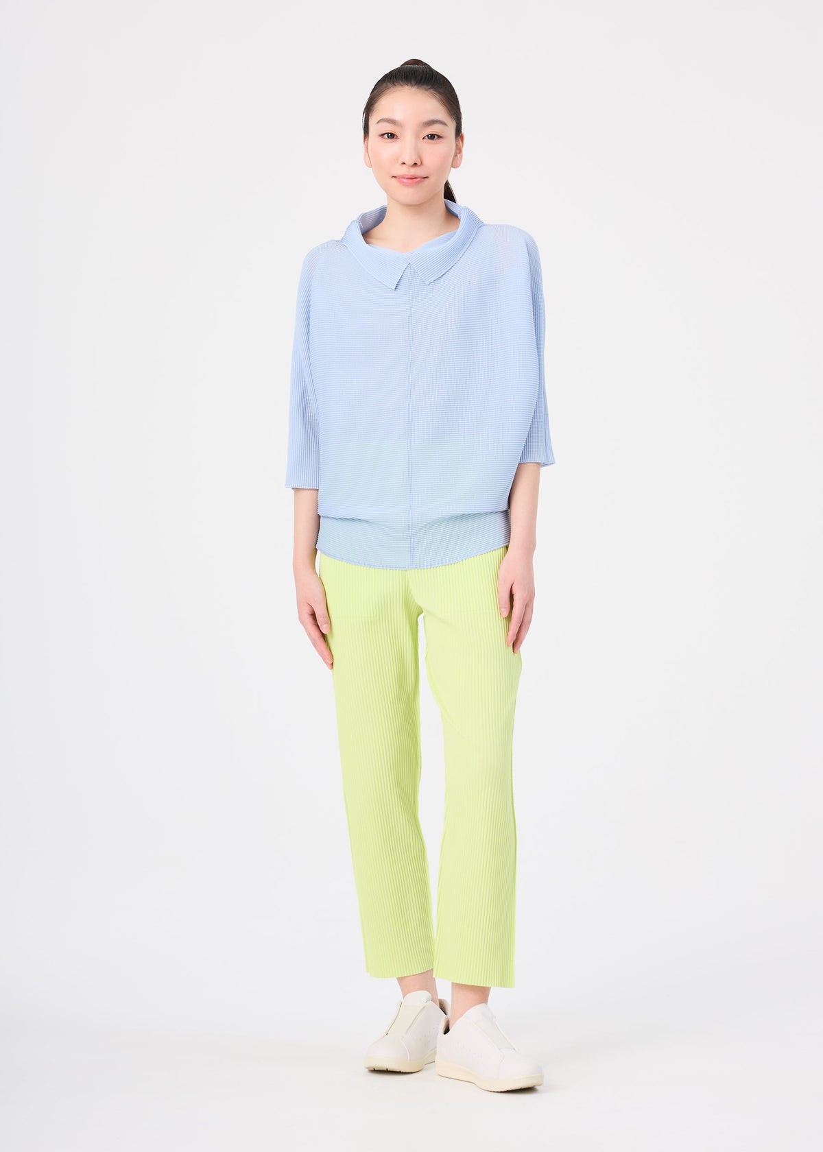 A woman faces forward against a white background, wearing me ISSEY MIYAKE's FINE KNIT PLEATS COLOR 1—a lightweight light blue top with three-quarter sleeves—paired with pale yellow straight pants and white slip-on shoes. view 6 of 7