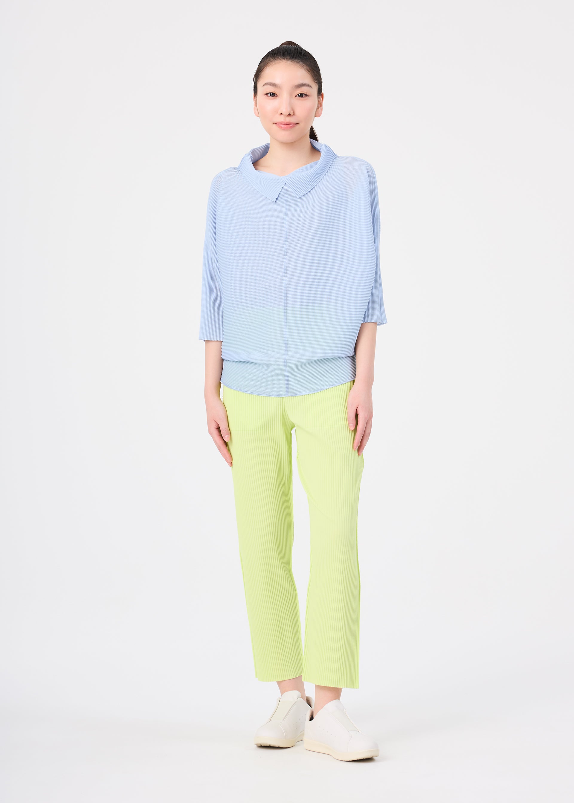 Zoomed view A woman faces forward against a white background, wearing me ISSEY MIYAKE's FINE KNIT PLEATS COLOR 1—a lightweight light blue top with three-quarter sleeves—paired with pale yellow straight pants and white slip-on shoes.