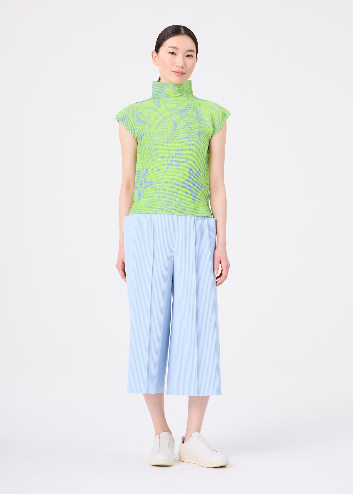 A woman with straight dark hair pulled back wears the me ISSEY MIYAKE FINE KNIT PLEATS COLOR 1 light blue wide pants, a green patterned sleeveless turtleneck top, and white slip-on shoes, standing against a white background. view 6 of 7