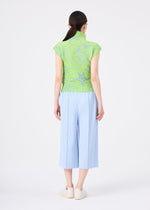 A woman with dark hair in a low ponytail faces away, wearing me ISSEY MIYAKE’s FINE KNIT PLEATS COLOR 1 wide light blue pants and a light green patterned top, styled with white slip-on shoes against a plain white background.