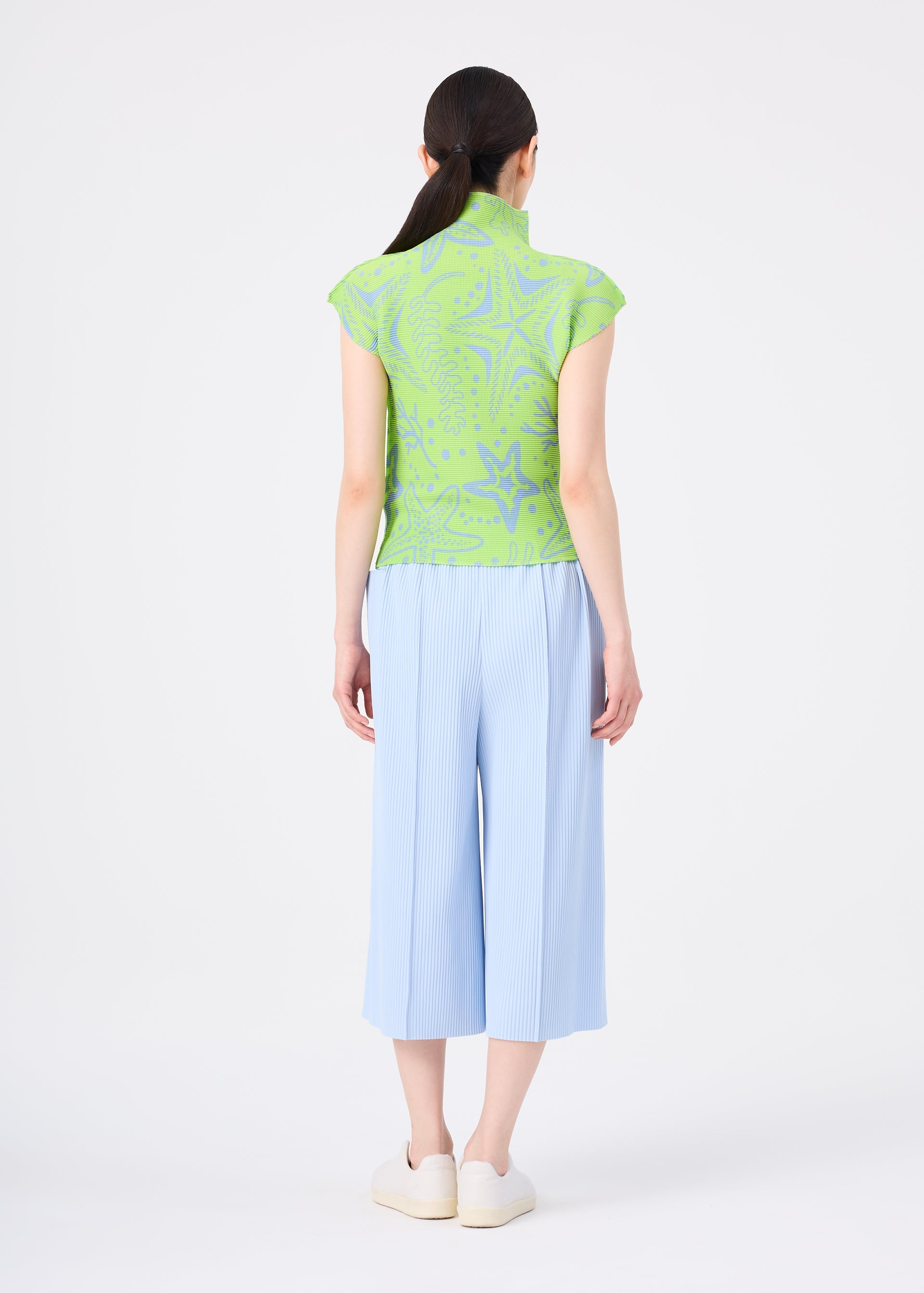 Zoomed view A woman with dark hair in a low ponytail faces away, wearing me ISSEY MIYAKE’s FINE KNIT PLEATS COLOR 1 wide light blue pants and a light green patterned top, styled with white slip-on shoes against a plain white background.