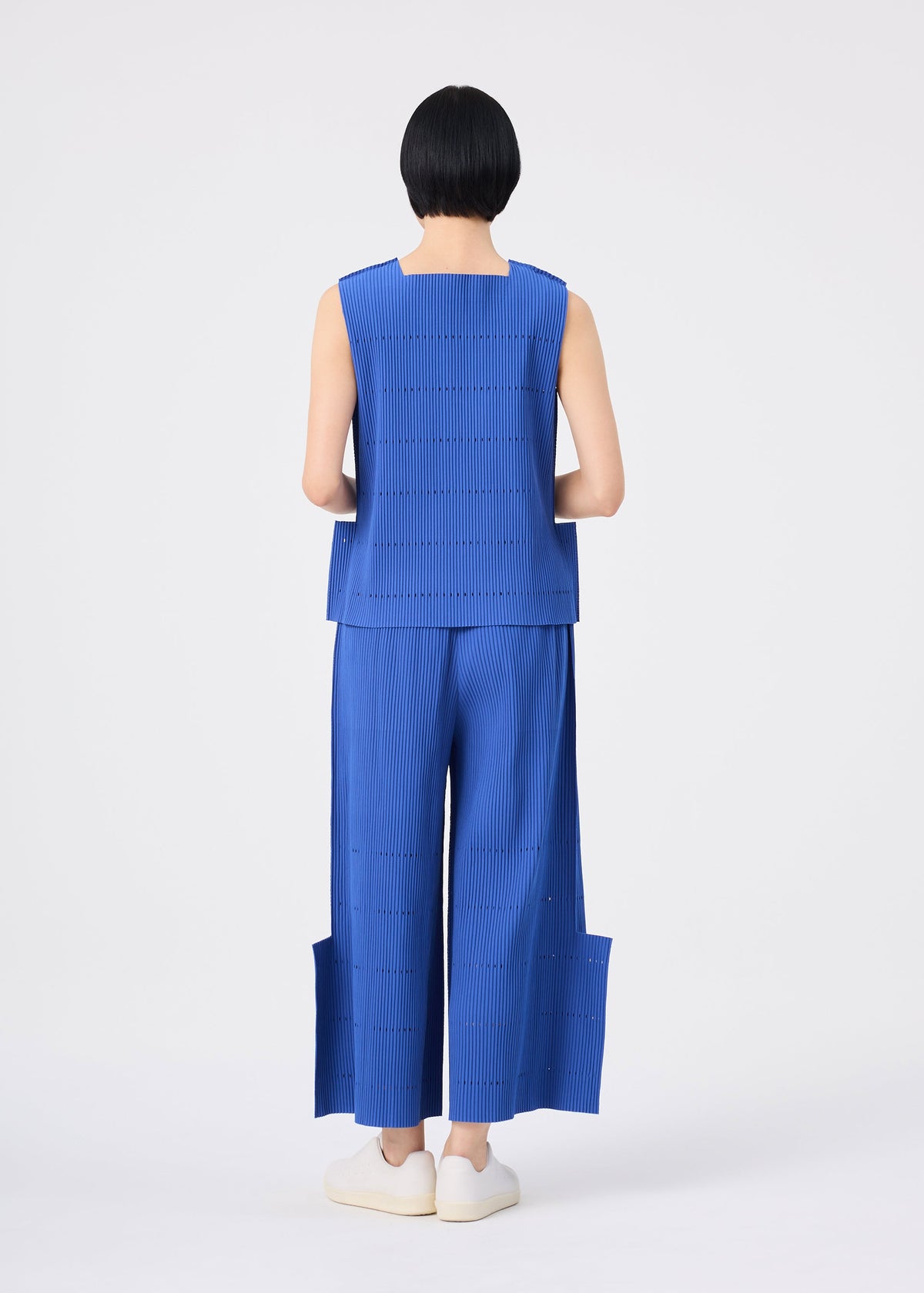 A person with short black hair stands facing away against a plain white background, wearing a sleeveless blue top and the FINE KNIT PLEATS BOTTOM 1 by me ISSEY MIYAKE—wide pleated knit pants featuring unique side panels—paired with white slip-on shoes. view 6 of 7