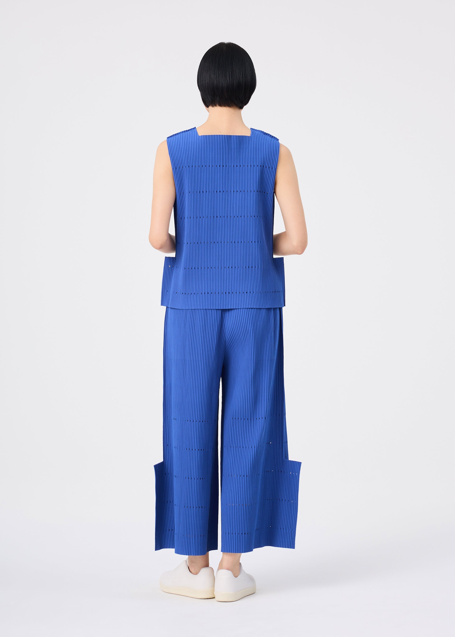 Zoomed view A person with short black hair stands facing away against a plain white background, wearing a sleeveless blue top and the FINE KNIT PLEATS BOTTOM 1 by me ISSEY MIYAKE—wide pleated knit pants featuring unique side panels—paired with white slip-on shoes.
