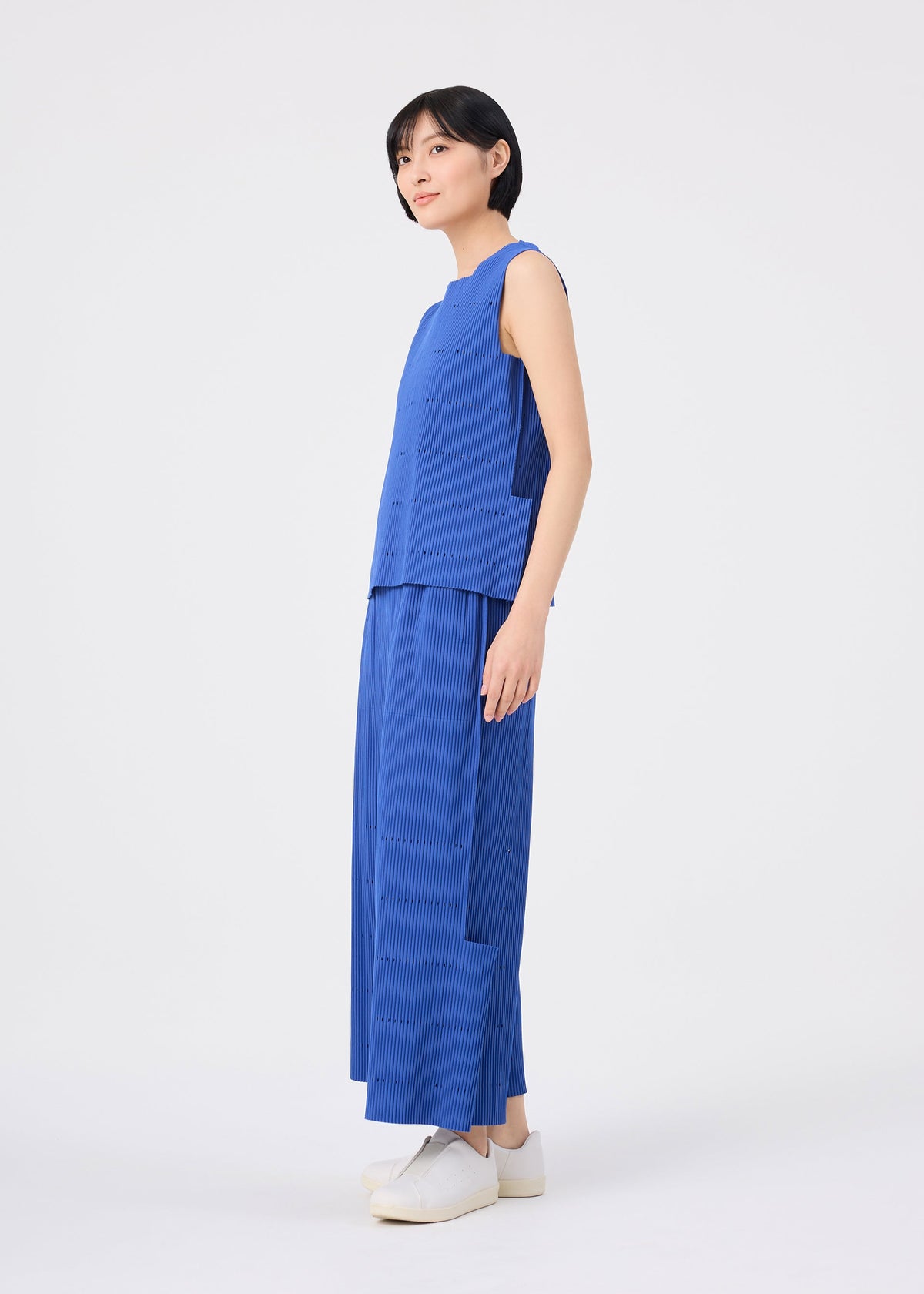A person stands sideways against a plain white background, wearing the me ISSEY MIYAKE FINE KNIT PLEATS BOTTOM 1—wide, pleated knit pants in blue—paired with a sleeveless blue top and white sneakers. view 7 of 7