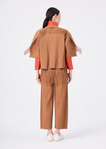 A woman stands facing away against a plain white background, wearing the me ISSEY MIYAKE LEAF PLEATS brown pleated knit pants featuring fringe details on the hem, an orange top underneath, and white shoes.