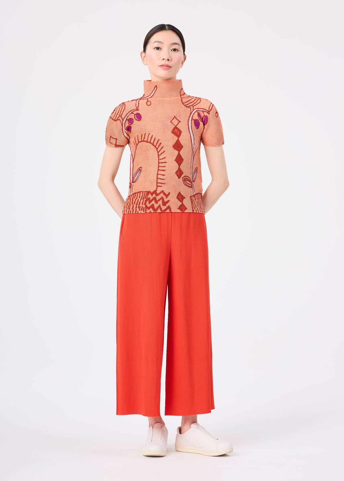 A person stands against a plain background wearing the FINE KNIT PLEATS COLOR 2 wide-legged red pants from me ISSEY MIYAKE, crafted from soft knit material. They have their hands behind their back and are paired with a peach patterned top and white slip-on shoes. view 6 of 7