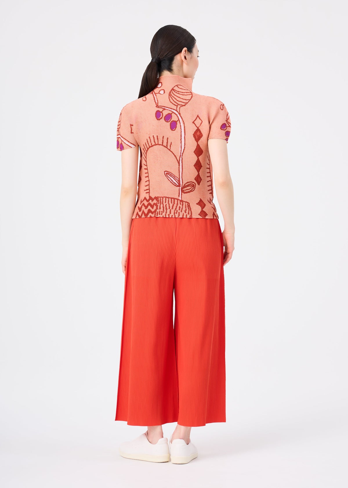 A woman stands facing away, dressed in bright red FINE KNIT PLEATS COLOR 2 pants by me ISSEY MIYAKE, featuring fine knit pleats. She pairs them with a peach short-sleeve top adorned with a bold abstract design. Her dark hair is tied back, she wears white shoes, and the background is plain white. view 7 of 7