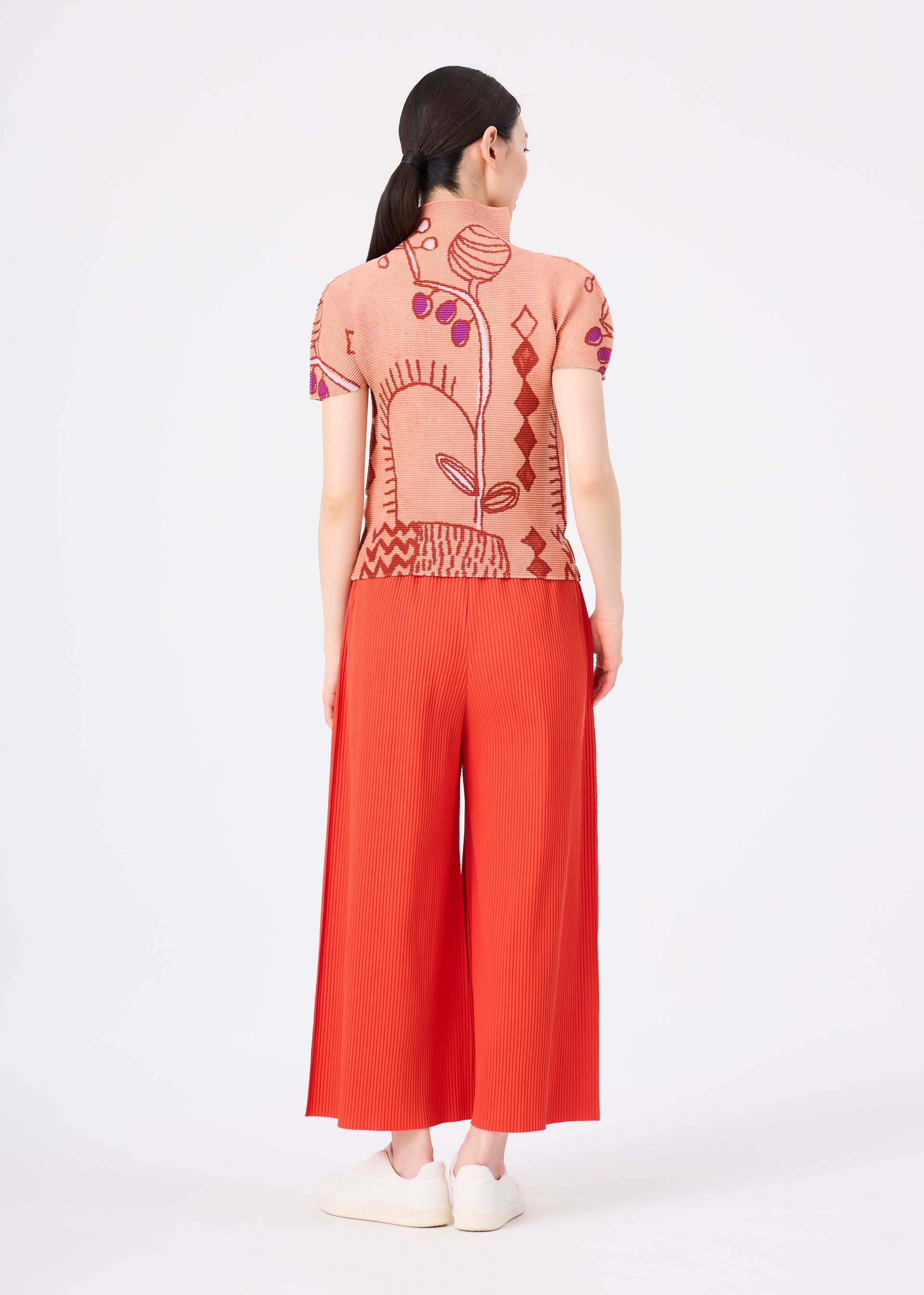 Zoomed view A woman stands facing away, dressed in bright red FINE KNIT PLEATS COLOR 2 pants by me ISSEY MIYAKE, featuring fine knit pleats. She pairs them with a peach short-sleeve top adorned with a bold abstract design. Her dark hair is tied back, she wears white shoes, and the background is plain white.