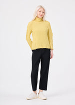 A person with long light hair wears a yellow textured turtleneck, MIX FINE KNIT PLEATS BOTTOM by me ISSEY MIYAKE, and white sneakers, standing against a plain white background.