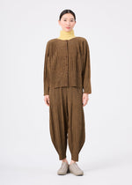 A woman faces forward against a white background, wearing a loose brown button-up top, the MIX FINE KNIT PLEATS BOTTOM by me ISSEY MIYAKE, a light yellow turtleneck underneath, and light beige shoes.