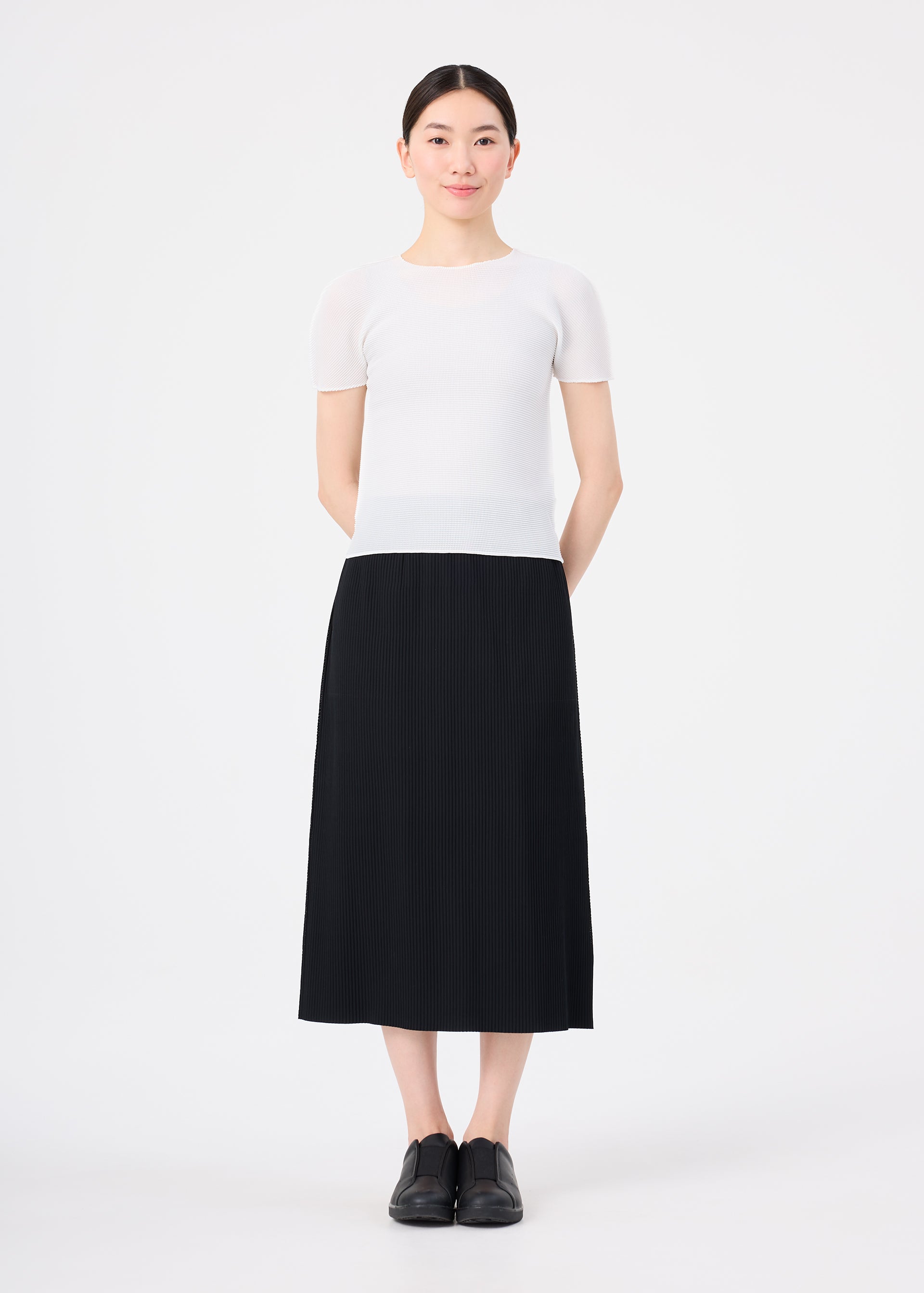 Zoomed view A woman stands against a white background with her hands behind her back, wearing the FINE KNIT PLEATS skirt by me ISSEY MIYAKE, a short-sleeved white top, and black shoes, maintaining a neutral expression.