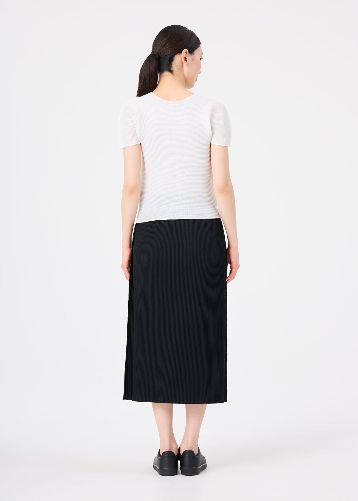 A woman stands facing away, wearing me ISSEY MIYAKE's FINE KNIT PLEATS black skirt with a short-sleeved white top and black shoes. Her dark hair is tied back in a low ponytail against a plain white background. view 6 of 7