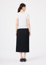 A woman stands facing away, wearing me ISSEY MIYAKE's FINE KNIT PLEATS black skirt with a short-sleeved white top and black shoes. Her dark hair is tied back in a low ponytail against a plain white background.