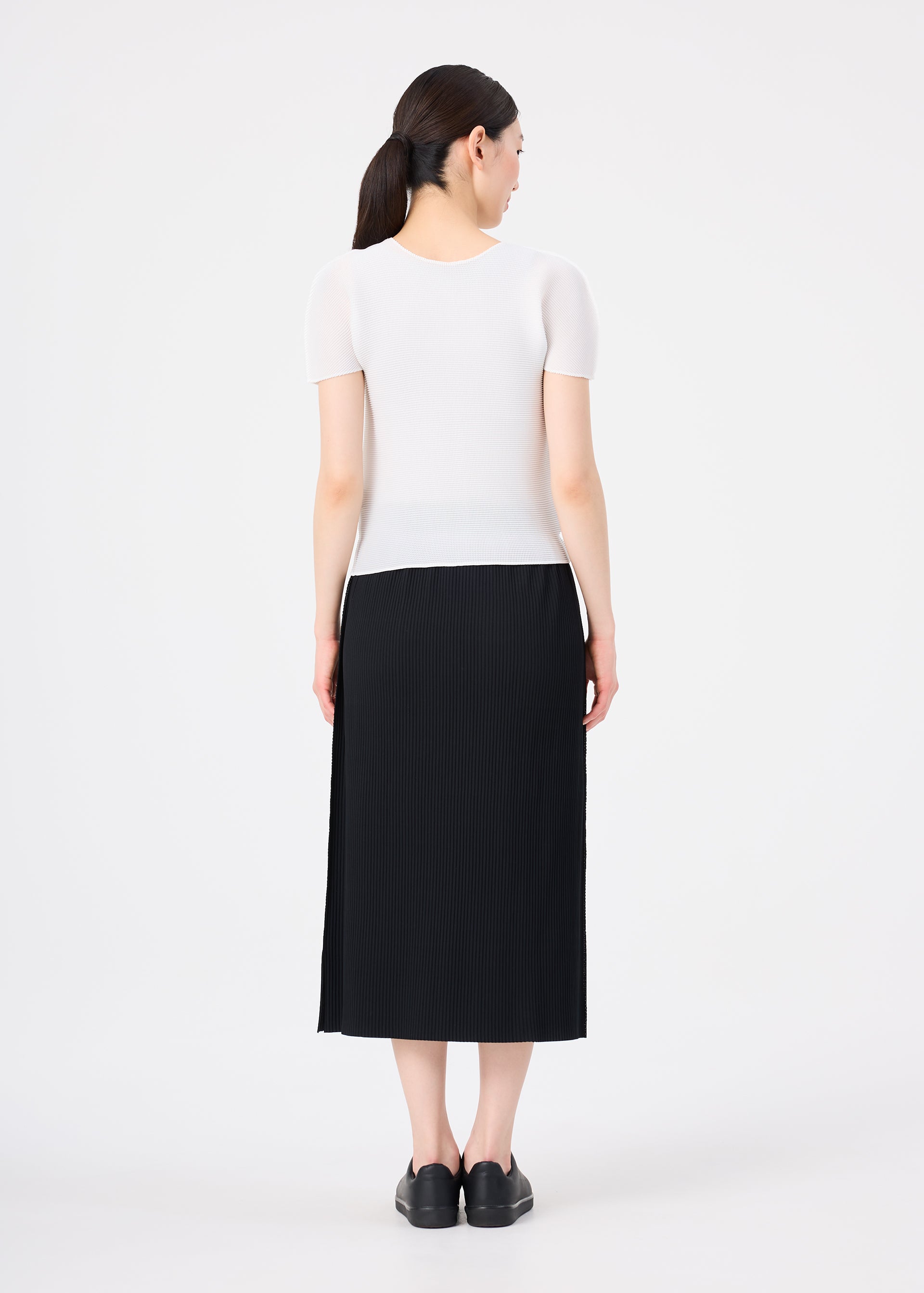 Zoomed view A woman stands facing away, wearing me ISSEY MIYAKE's FINE KNIT PLEATS black skirt with a short-sleeved white top and black shoes. Her dark hair is tied back in a low ponytail against a plain white background.