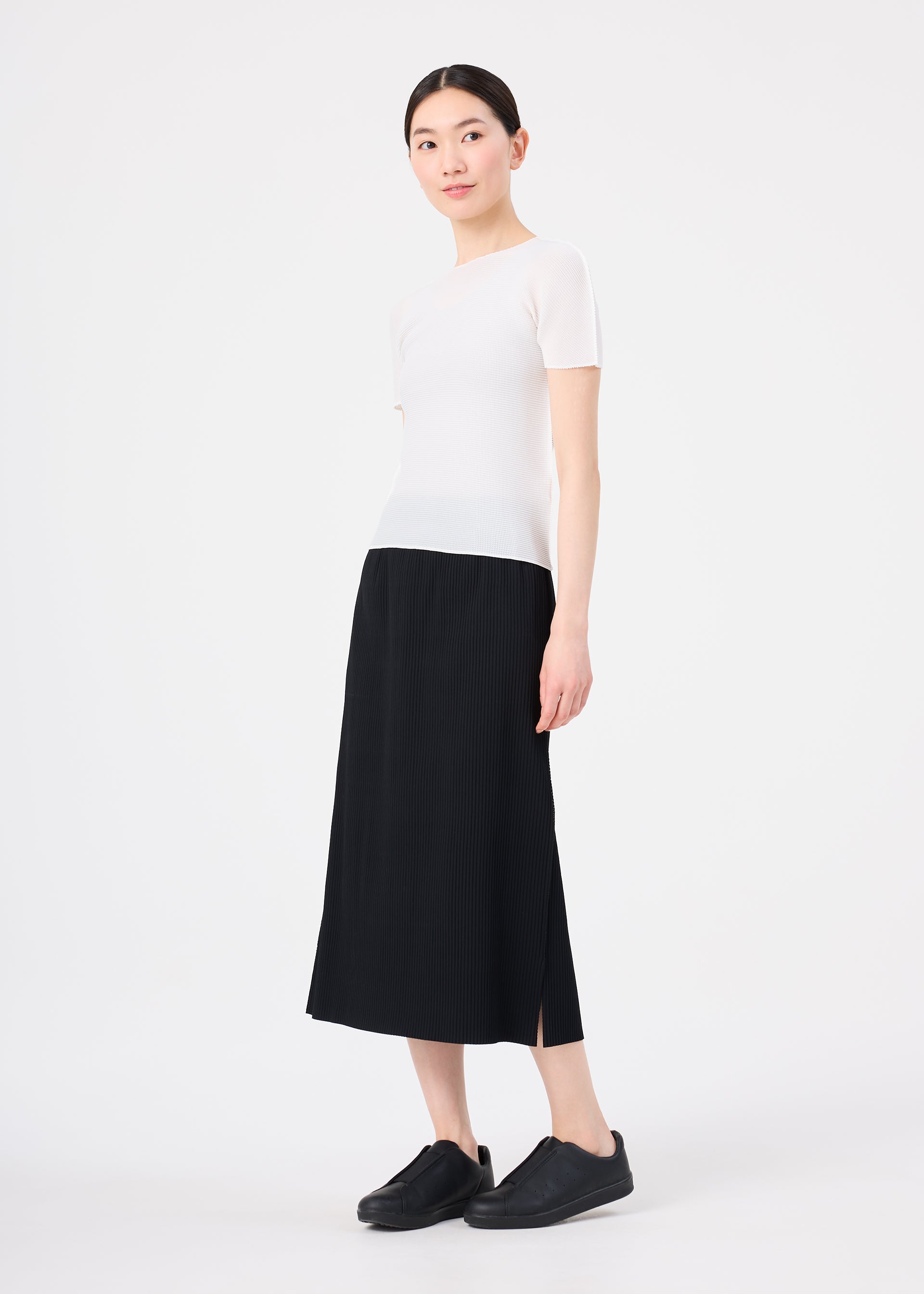 Zoomed view Against a plain white background, a person with tied-back black hair and a slight smile wears me ISSEY MIYAKE’s FINE KNIT PLEATS black pleated skirt paired with a short-sleeved white top and black slip-on shoes.