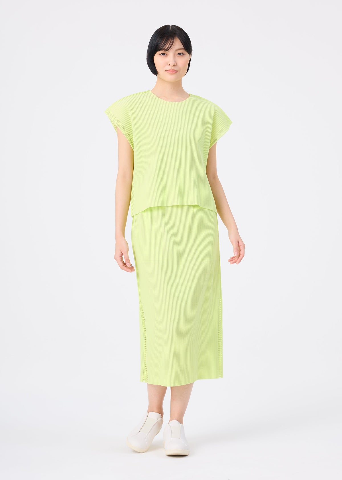 A person stands against a plain white background, wearing the MESH PLEATS short-sleeve top and matching pleated skirt by me ISSEY MIYAKE in light green, paired with white slip-on shoes. view 5 of 7