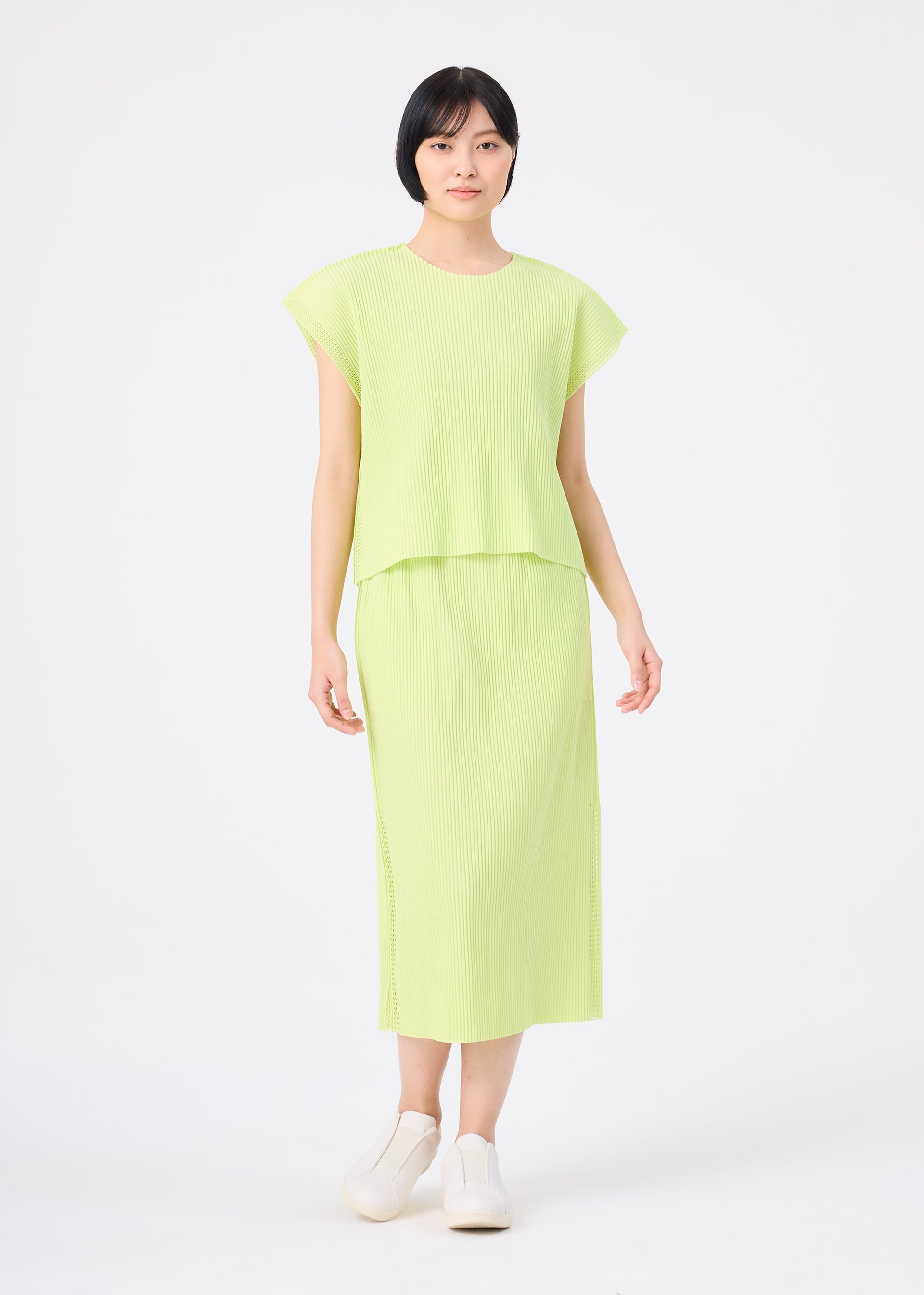 Zoomed view A person stands against a plain white background, wearing the MESH PLEATS short-sleeve top and matching pleated skirt by me ISSEY MIYAKE in light green, paired with white slip-on shoes.