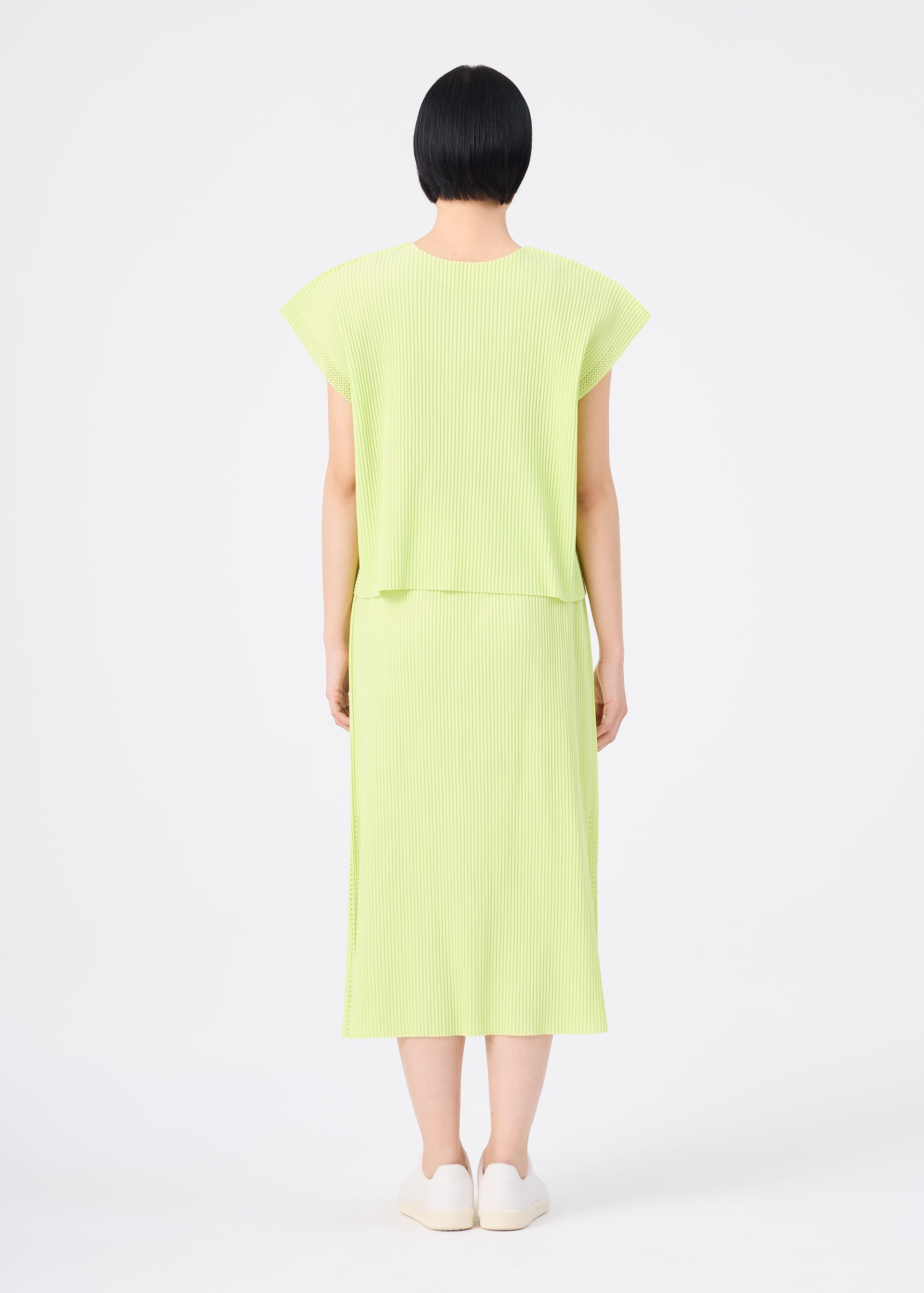 Zoomed view A person with short black hair is standing facing away, wearing the me ISSEY MIYAKE MESH PLEATS light green, short-sleeved, knee-length pleated knit skirt dress and white slip-on shoes, against a plain white background.