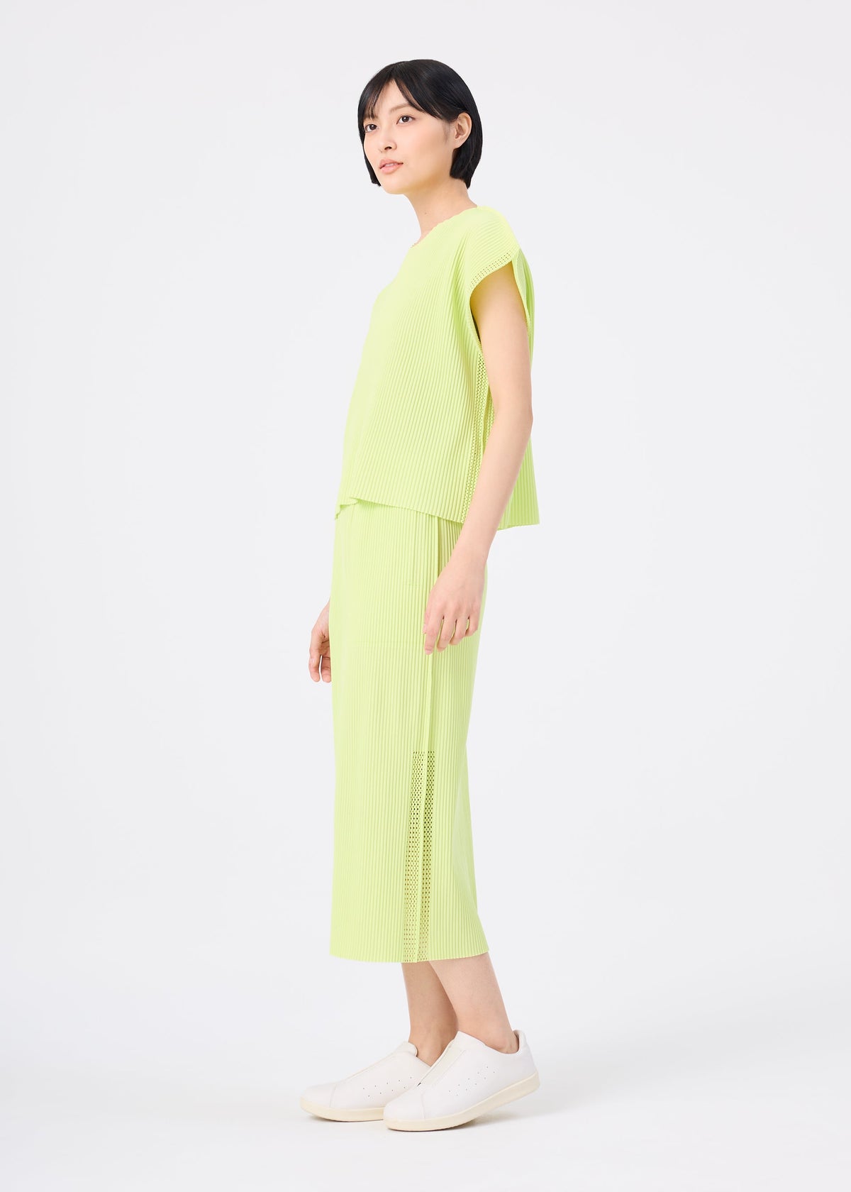 A woman with short black hair stands sideways against a plain white background, wearing the light green MESH PLEATS set from me ISSEY MIYAKE paired with white sneakers. view 7 of 7