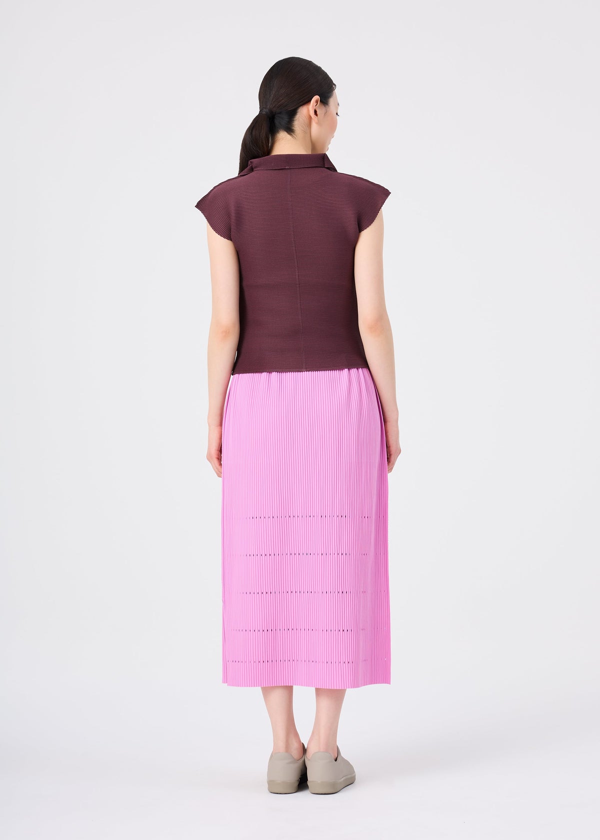 A woman stands facing away, wearing a short-sleeve maroon top and the FINE KNIT PLEATS BOTTOM 1 skirt by me ISSEY MIYAKE in pink, paired with beige shoes. The background is plain white. view 7 of 8
