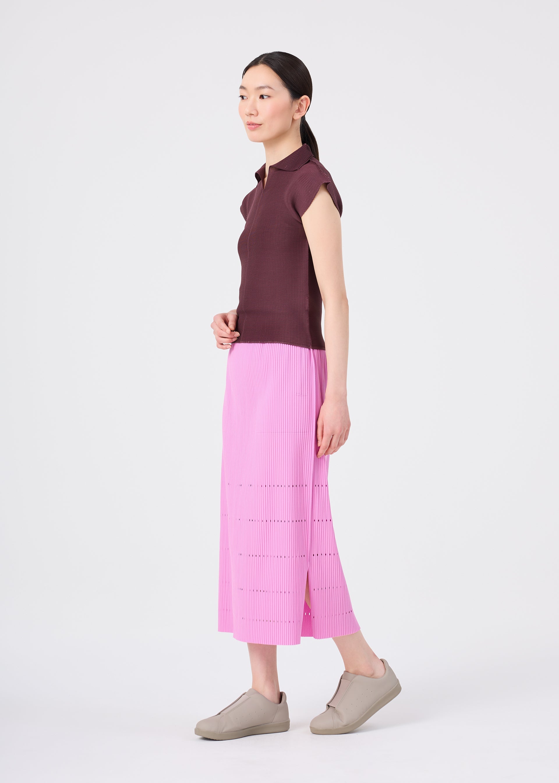 Zoomed view A woman with a ponytail stands sideways against a plain white background, wearing a short-sleeve brown top, light gray slip-on shoes, and the FINE KNIT PLEATS BOTTOM 1 pink pleated knit skirt from me ISSEY MIYAKE.