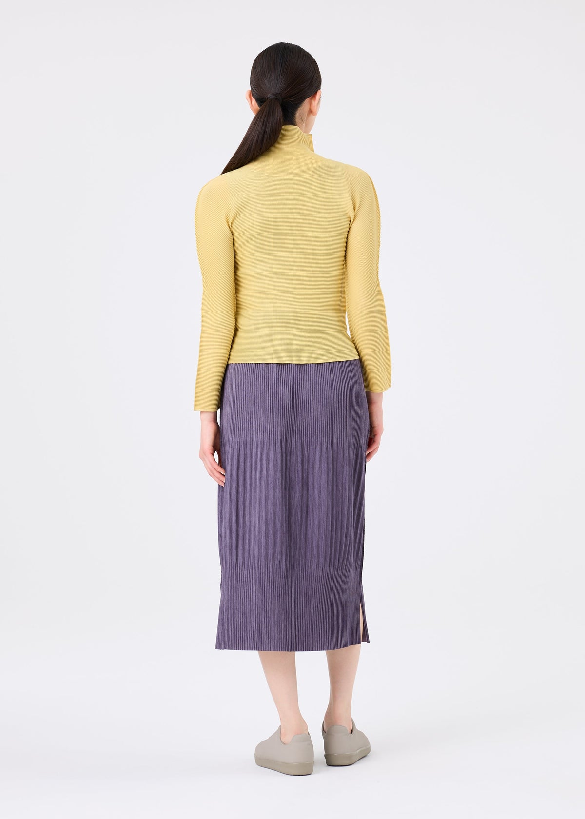 A woman stands with her back to the camera, wearing a yellow long-sleeve top and the MIX FINE KNIT PLEATS BOTTOM by me ISSEY MIYAKE. She pairs beige shoes with her low ponytail against a plain white background. view 8 of 8