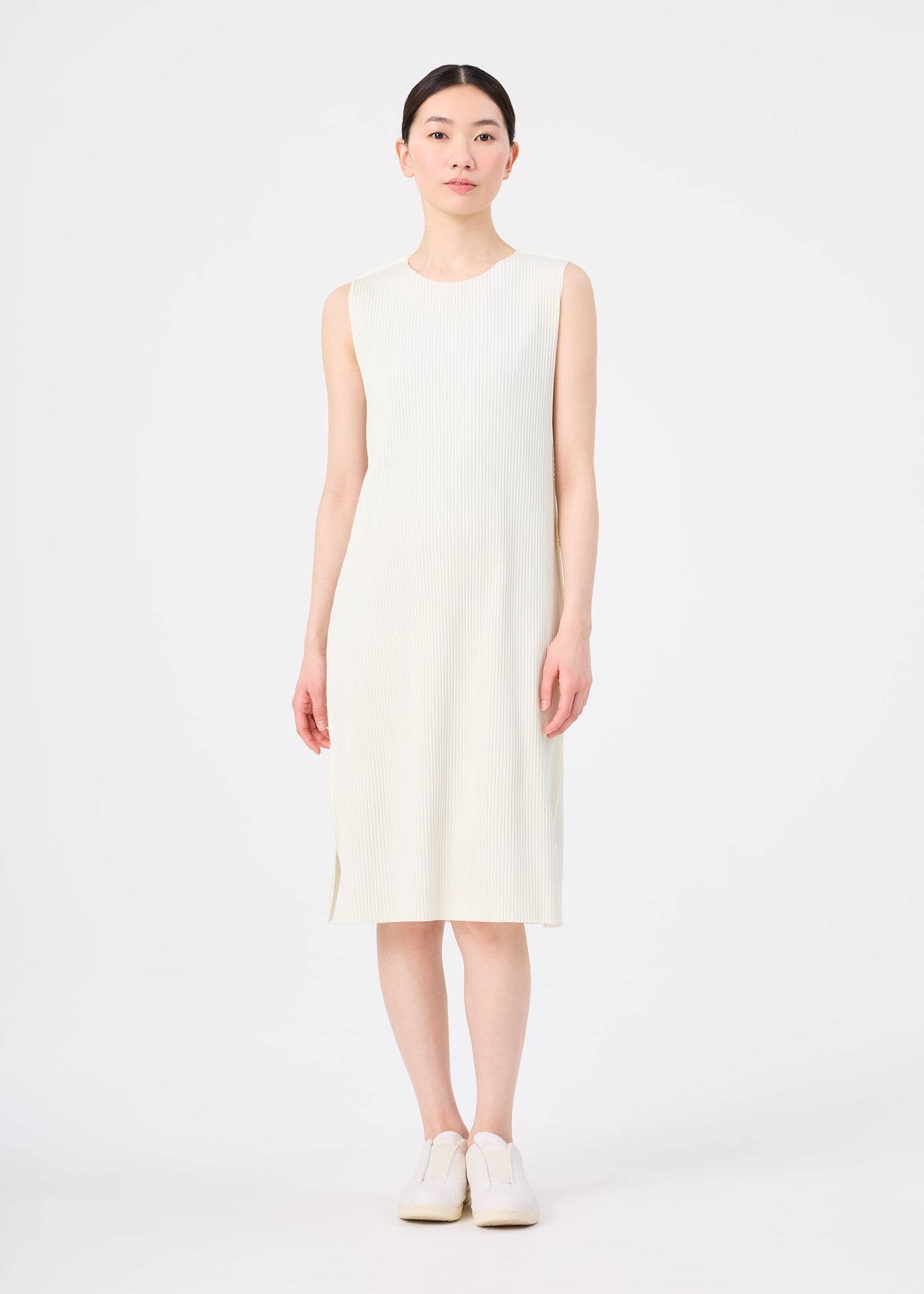 A woman faces forward against a white background, wearing the me ISSEY MIYAKE FINE KNIT PLEATS sleeveless ivory knee-length dress with fine knit pleats and white shoes, arms relaxed by her sides. view 5 of 7