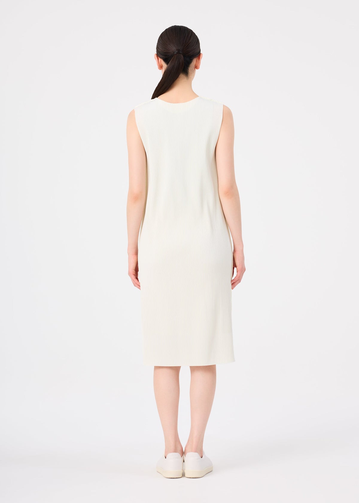 A woman with dark hair in a low ponytail stands facing away, wearing a sleeveless, knee-length cream FINE KNIT PLEATS dress from me ISSEY MIYAKE and white slip-on shoes against a plain white background. view 6 of 7